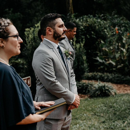 Lily, wearing a navy blue dress, stands with the groom at a wedding. The groom is a light-skinned Latino man with a short beard and is wearing a light gray suit.
