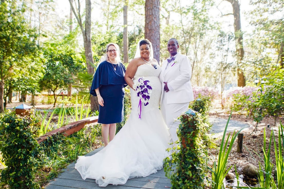 A Black woman with medium skin wearing a chapel length wedding dress and her wife, a Black woman with dark skin wearing a white tuxedo and purple bow tie, standing on a small wooden bridge. Lily stands to the left of them in a navy blue dress.